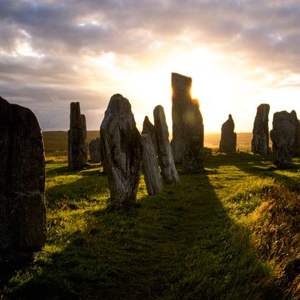 Ein prähistorischer Steinkreis oder eine Reihe von aufrechten Steinen in einer weiten, grasbewachsenen Landschaft bei Sonnenaufgang. Die Sonne scheint durch die Steine und wirft lange Schatten.