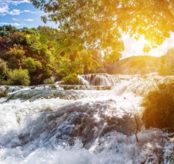 Eine Landschaft mit einem Wasserfall und Gebäuden bei Sonnenuntergang.