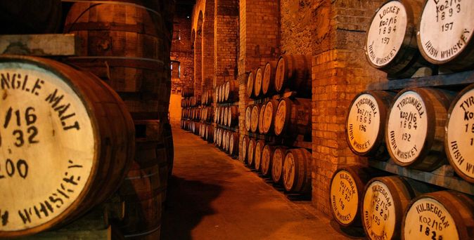 Ein beleuchteter Lagerraum mit Reihen von großen, hölzernen Whiskeyfässern, die auf Holzregalen gelagert sind. Viele Fässer sind mit handgeschriebenen Etiketten versehen.