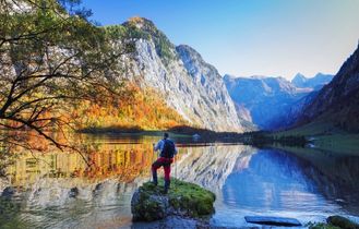 Eine Person steht auf einem Felsen im Königssee, Berchtesgaden, umgeben von herbstlichen Bergen und spiegelndem Wasser.