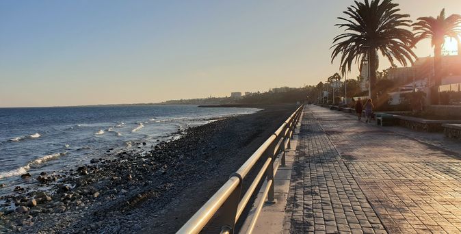 asphaltierte Promenade, links daneben Sandstrand und das blazue Meer, Hintergrund Palme, blauer Himmel kurz nach Sonnenuntergang