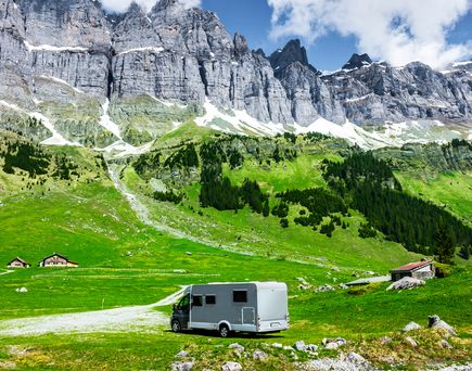 Ein weißer Camp am Ufer eines Sees, ein Wiesenweg führt zu der Bergkette im Hintergrund.