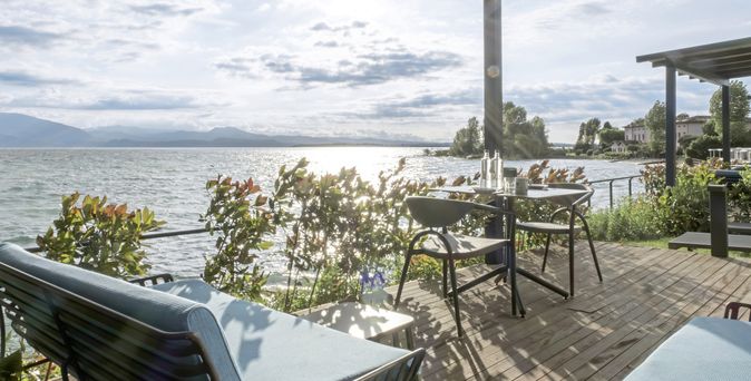 Terrasse mit Lounge-Möbeln und Esstisch, Blick auf einen See bei tiefstehender Sonne unter schattenspendender Pergola.