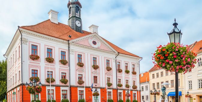 Rathaus in Tartu, davor der Marktplatz und eine Laterne mit Blumen unterhalb der Lampe. Das Rathaus ist orange und weiß gehalten und erstreckt sich über zwei Gebäude.