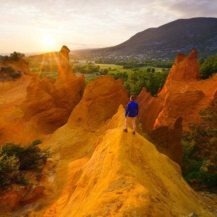 Eine weite Landschaft mit rötlichen Hügeln und Tälern, die von der untergehenden Sonne in warmes Licht getaucht werden. Die Szene wirkt sanft und malerisch.