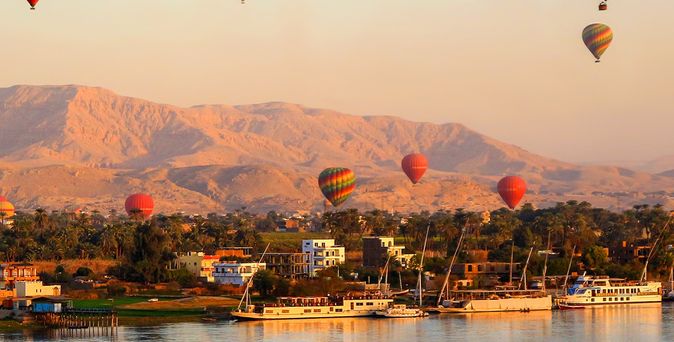 Mehrere bunte Heißluftballons schweben über den antiken Tempeln und der Wüstenlandschaft von Luxor, Ägypten, bei Sonnenaufgang.