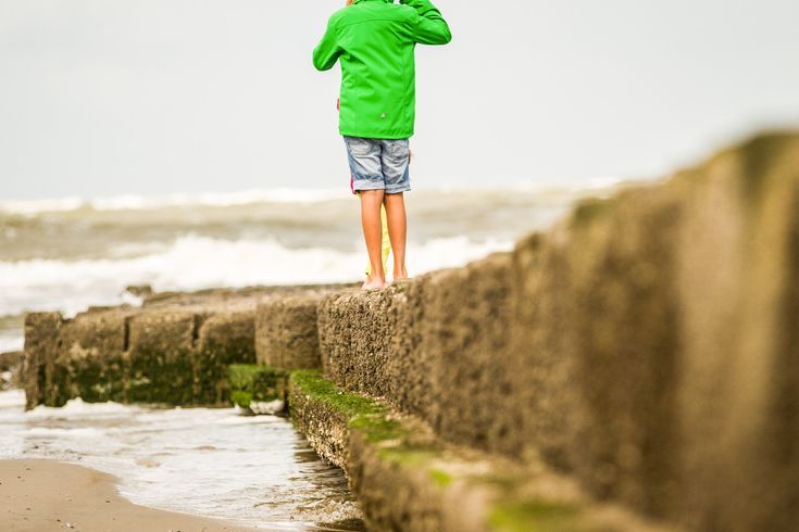 Ein Kind mit grüner Jacke und kurzer Hose spaziert auf einem Steinsteg am Strand.