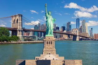 Die Freiheitsstatue ragt prominent aus dem Wasser auf, mit der Brooklyn Bridge und der beeindruckenden Skyline von Lower Manhattan im Hintergrund an einem klaren Tag.