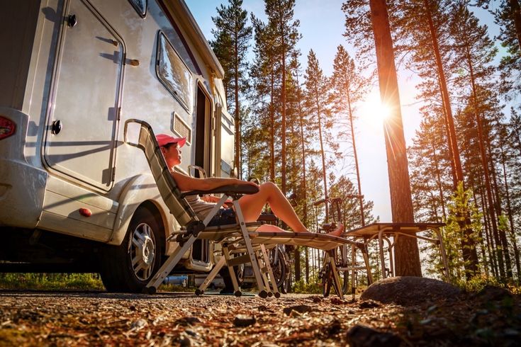 Ein Campervan parkt in einem sonnigen Wald, daneben ein Zelt und Campingmöbel.