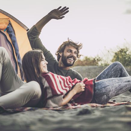 Ein Paar entspannt vor einem geöffneten Zelt auf einem Campingplatz, junge Frau blickt zum Mann.