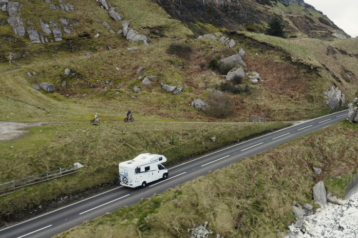Ein weißes Wohnmobil fährt auf einer kurvenreichen Straße durch eine weite, grasbewachsene Landschaft mit Seen und Bergen im Hintergrund. Der Himmel ist bewölkt und spiegelt sich im Wasser.