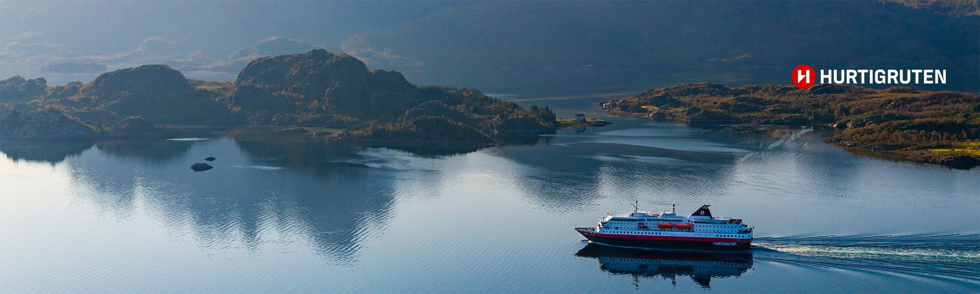 Luftaufnahme eines Hurtigruten-Schiffes, das durch einen schmalen Wasserweg fährt. Die umliegende Landschaft besteht aus steilen, bewaldeten Bergen, die sich im Wasser spiegeln. Das Hurtigruten-Logo ist oben rechts sichtbar.