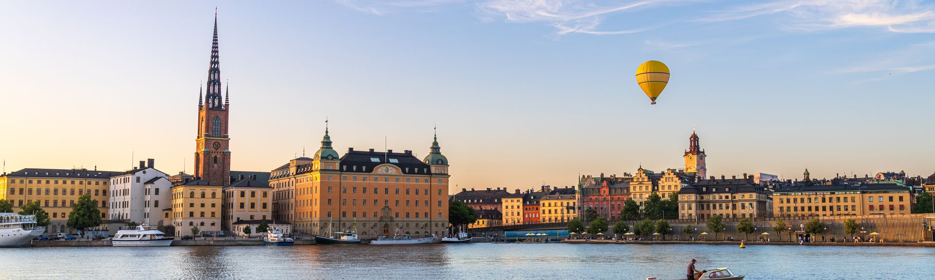 Blick auf die Silhouette von Riddarholmen gegen das Wasser, ein gelber Heißluftballon schwebt über den Gebäuden, Boot fährt vorbei, Stockholm, Schweden