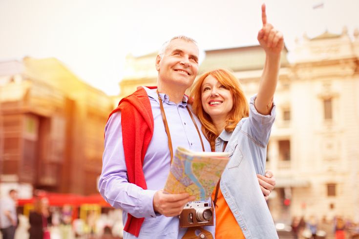 Fröhliches älteres Paar steht mit Stadtkarte und Kamera in der Stadt und schaut in die Ferne, die Frau zeigt mit dem Finger auf ein Ziel.
