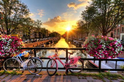 Bunte Fährrader über einer Gracht, am Geländer angestellt. Am Geländer einige Blumenkästen mit farbenfrohen Blumen. Das alles bei blauem Himmel bei Sonnenaufgang.