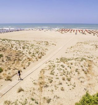 Breiter Sandstrand mit Holzstegzugang durch die Dünenlandschaft bis zu den Liegeplätzen mit Sonnenschirmen am Meer.