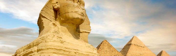 Eine Nahaufnahme der Großen Sphinx von Gizeh, mit zwei der Pyramiden von Gizeh im Hintergrund unter einem bewölkten Himmel.