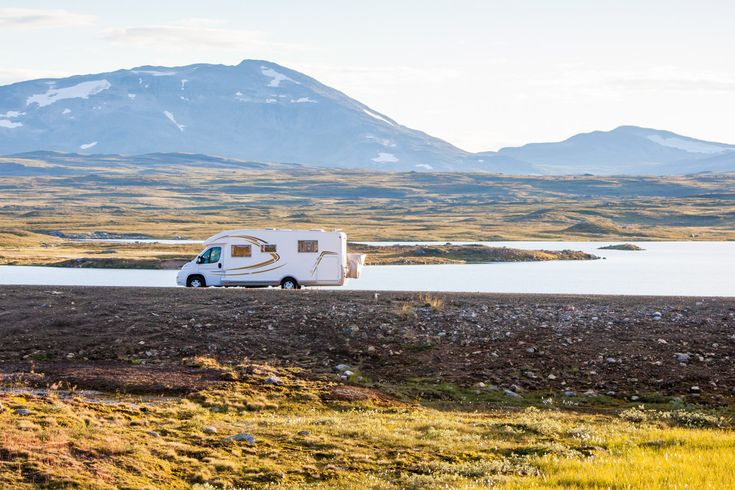 Weißer Campervan fährt auf einem Dammweg, mit Wasser auf der linken und Gras auf der rechten Seite, Berge im Hintergrund.