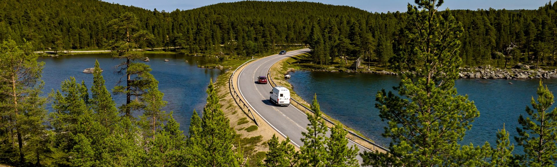 Wohnmobil und Auto fahren entlang einer Straße, welche von Wäldern und Gewässer umgeben ist, in Finnland 