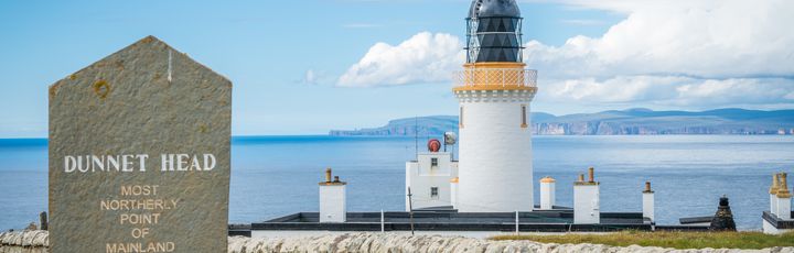 Blick auf Dunnet Head. In der Front steht ein Gedanken-/Meilenstein, dahinter ein weißer Leuchtturm zu sehen.