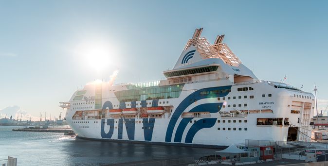 Ein großes weißes Fährschiff mit "GNV" in großen blauen Buchstaben an der Seite liegt bei Sonnenuntergang in einem Hafen. Das Schiff hat eine große, geschwungene blaue und weiße Struktur auf dem Oberdeck.