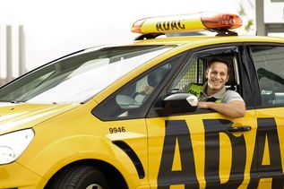 Freundlicher ADAC Mitarbeiter in Uniform sitzt am Steuer eines gelben ADAC Pannenhilfe-Fahrzeugs und lächelt aus dem Fenster.