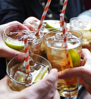 Vier Gläser mit Whiskey auf einem Holztisch, die auf einer dunklen Holzplatte stehen. Im Hintergrund sitzen zwei Personen an einem Tisch, deren Gesichter unscharf sind.