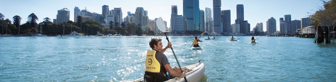 Ein Mann in einem gelben Kajak paddelt auf dem Brisbane River, während im Hintergrund die Skyline der Stadt Brisbane unter einem klaren blauen Himmel zu sehen ist. Weitere Kajakfahrer sind ebenfalls auf dem Wasser.