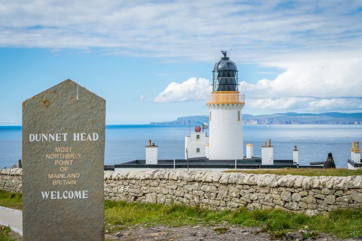 Blick auf Dunnet Head. In der Front steht ein Gedanken-/Meilenstein, dahinter ein weißer Leuchtturm zu sehen.